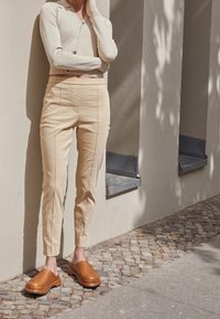 Une personne s'appuie contre un mur beige, vêtue d'un cardigan crème, d'un pantalon taille haute beige et de chaussures à enfiler marron sur un pavé en pierre.