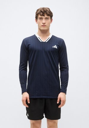 Marineblaues langärmliges Sportshirt mit vertikalen Streifen, V-Ausschnitt und weißem Akzent am Kragen; kombiniert mit schwarzen Shorts mit Logo.