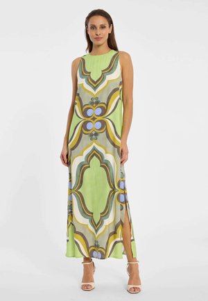 Frau trägt ein ärmelloses, knöchellanges Kleid mit grünen, braunen, weißen, gelben und blauen geometrischen Mustern und einem seitlichen Schlitz, kombiniert mit beigen High Heels.