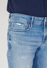 Vaqueros de mezclilla azul claro con una textura desgastada, bolsillos funcionales y una pequeña etiqueta en la parte posterior. Detalles desgastados visibles.