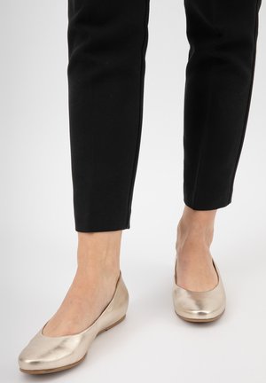 Ballerine dorate metalliche con una superficie liscia e punta arrotondata, indossate con pantaloni neri attillati. Suola piatta e design semplice.