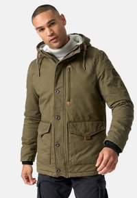 Olijfgroene parka met een capuchon, gestructureerde stof, voorknopsluiting, twee grote zakken en leren accenten. Gelaagd over een gebreide trui.