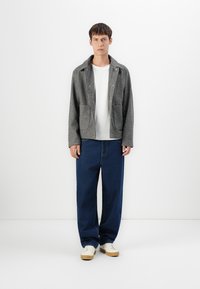 Ung mann står mot hvit vegg iført grå zip-up jakke over hvit t-skjorte, løse blå jeans og hvite joggesko.