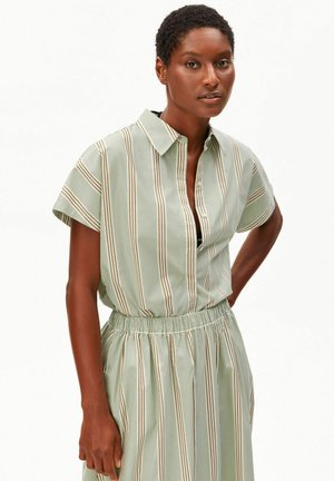 Frau trägt ein salbeigrünes, kurzärmeliges, gestreiftes Hemdkleid mit Kragen und elastischem Bund, steht mit einer Hand in der Hüfte vor weißem Hintergrund.