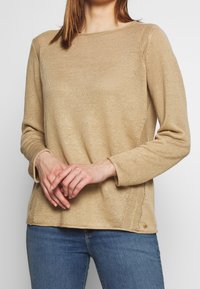 Beige gestrickter Pullover mit Rundhalsausschnitt, langen Ärmeln und strukturierter Oberfläche. Besitzt seitliche Schlitze und einen kleinen Akzent am Saum.