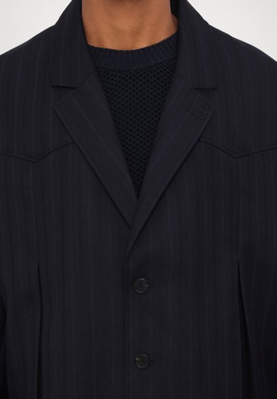 Marineblauer Nadelstreifen-Blazer mit Revers, zwei Knopfschluss und weicher Textur, über einem marineblauen Strickpullover getragen.