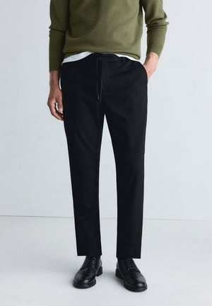 Homme portant un pantalon noir sur mesure, des chaussures habillées noires, un pull vert olive et un sous-vêtement blanc, debout devant un fond clair uni.