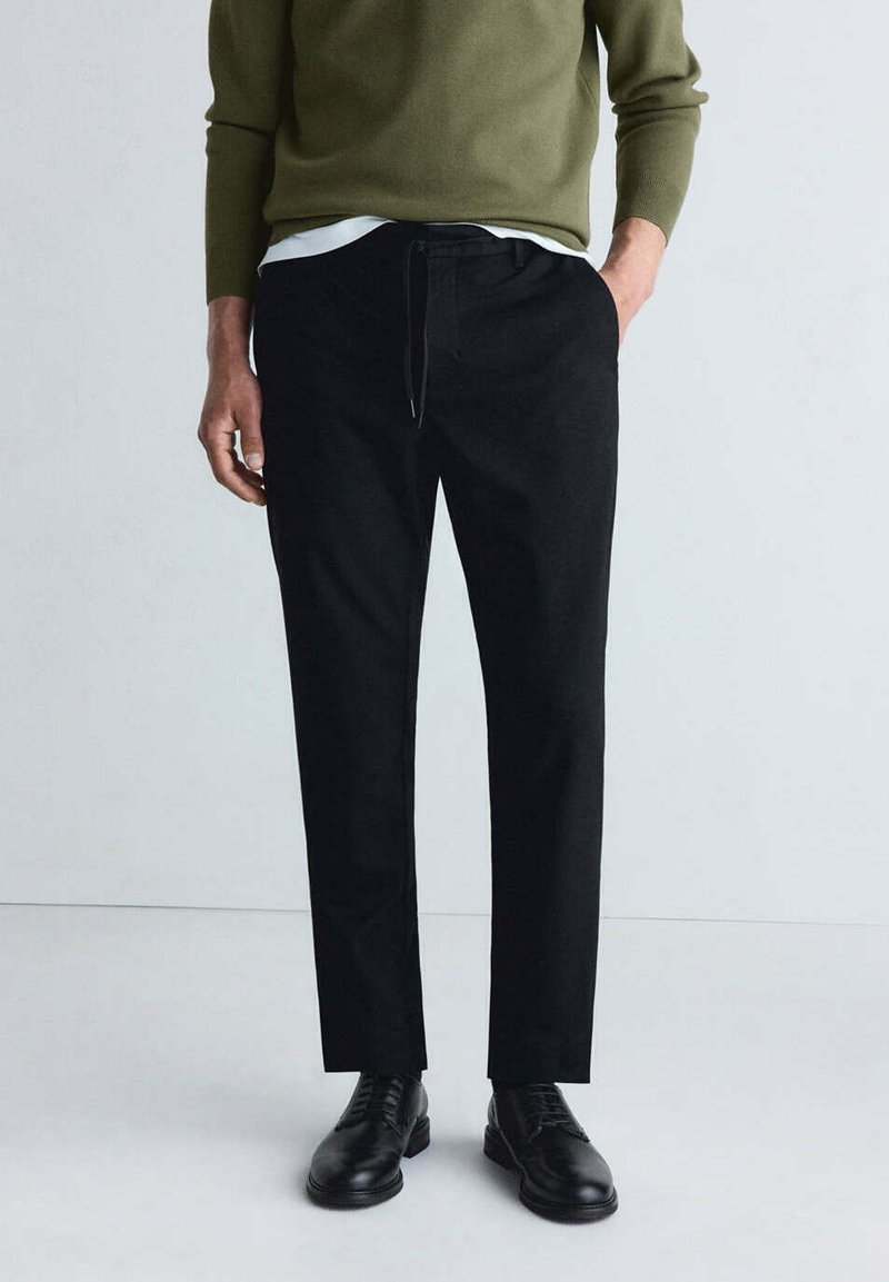 Homme portant un pantalon noir sur mesure, des chaussures habillées noires, un pull vert olive et un sous-vêtement blanc, debout devant un fond clair uni.