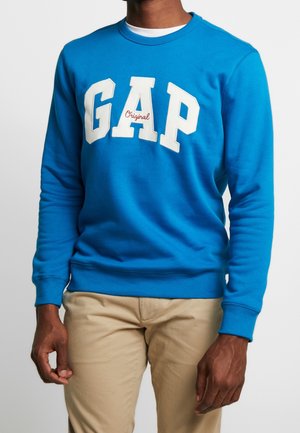Mężczyzna noszący jasnoniebieską bluzę z napisem „GAP Original” w białym i czerwonym na wysokości klatki piersiowej, w połączeniu z beżowymi spodniami.