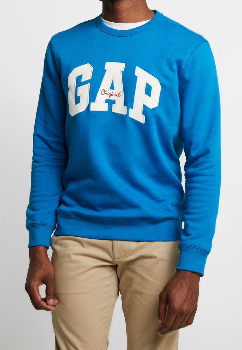 Homme portant un sweat-shirt bleu vif avec le texte « GAP Original » en blanc et rouge sur la poitrine, associé à un pantalon beige.