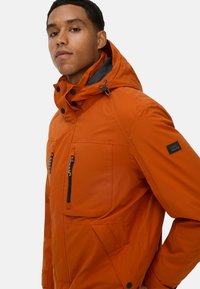 Pomarańczowa wodoodporna kurtka z kapturem, wyposażona w kieszenie na zamek błyskawiczny, regulowane sznurki oraz teksturowane wykończenie. Z dodatkiem naszywki z logo.