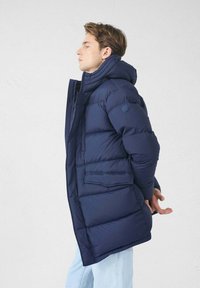 Un homme se tenant de côté, les yeux fermés, portant une veste matelassée bleu marine avec capuche et un jean bleu clair, sur un fond uni.