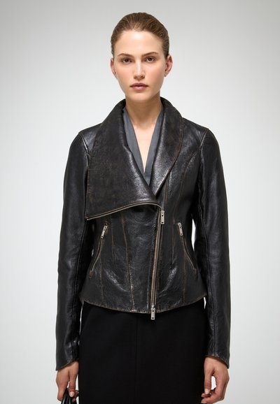 Nainen, joka pukeutunut mustaan epäsymmetriseen nahkatakkiin, jossa vetoketju, ja harmaan paidan alla, pitää mustaa laukkua yksinkertaisen taustan edessä.