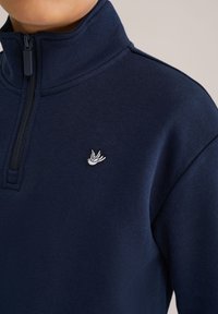 Temno modra pulover s polovičnim zadrgo in teksturirano površino, ki ima na prsih majhen beli vezen ptičji logotip in stojni ovratnik.