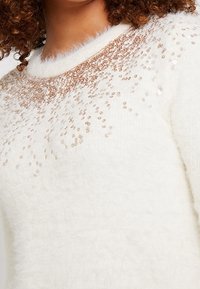 Valkoinen pörröinen neule, jossa on pyöreä alaosa ja joustava suunnittelu, jossa on paljettikuvio, joka vaihtelee olkapäiden ympärillä. Pehmeä ja teksturoitu materiaali.