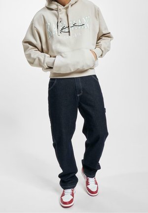 Persoon in een beige hoodie met de tekst "Karl Kani", een donkere spijkerbroek en rood-witte sneakers, staand met de handen in de zakken van de hoodie.