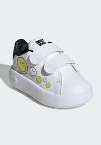 White sneakers with a smooth material featuring yellow smiley face designs. Velcro straps, rounded toe, and textured black lining.