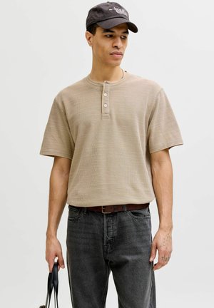Mann trägt beige kurzärmeliges Henley-Shirt und schwarze Kappe, hält schwarze Tragetasche und steht vor schlichtem hellgrauem Hintergrund.
