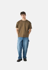 Brown cotton t-shirt with short sleeves, paired with light blue denim jeans. Model standing with hands behind, wearing black shoes.
