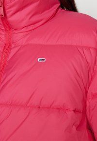 Nainen, jolla on yllään kirkkaan vaaleanpunainen untuvatakki, jossa on pieni suorakulmainen logo rinnassa ja näkyvä etuvetoketju.