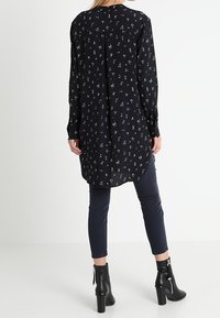 Svart tunika med liten blommönster, långa ärmar, rundad nederkant och detalj med plissering i ryggen, ihop med mörka jeans och svarta klackstövlar.