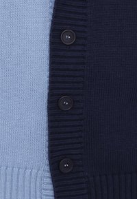 Cardigã de malha com um design bicolor, apresentando seções em azul claro e azul marinha. Seis botões pretos e uma bainha canelada proporcionam textura.