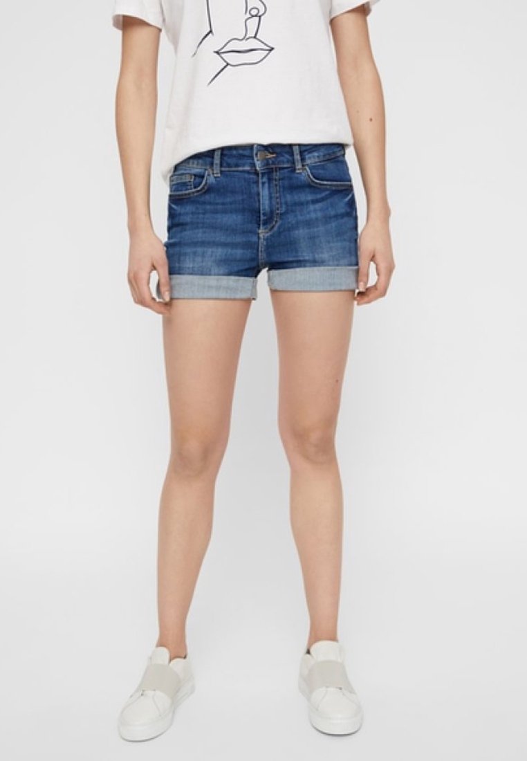 Personne portant un short en jean bleu retroussé, un t-shirt blanc avec un dessin de visage en ligne, et des baskets blanches sans lacets.