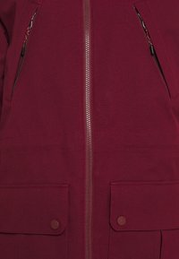 Giacca bordeaux con zip completa, due tasche frontali con chiusura a scatto e un tessuto testurizzato, con dettagli della zip nascosti.
