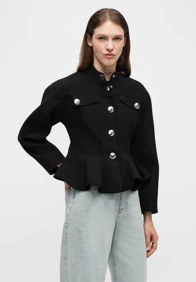 Γυναίκα που φοράει μαύρο εφαρμοστό σακάκι με μεγάλα ασημένια κουμπιά και ανοιχτό μπλε τζιν με ψηλή μέση, στέκεται με το ένα χέρι στην τσέπη.