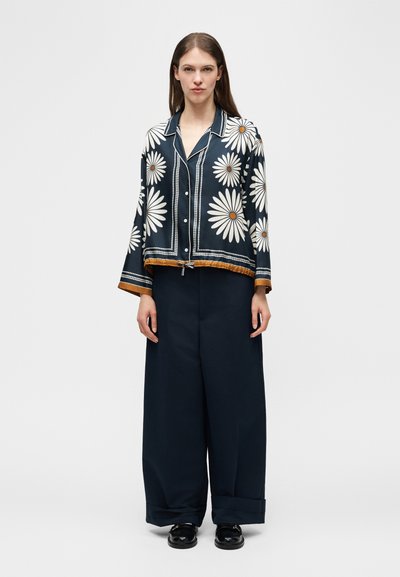 Γυναίκα που φοράει μπλε μπλούζα με μεγάλα λευκά μαργαρίτες και μαύρο παντελόνι με φαρδιά πόδια, στέκεται με τα χέρια χαλαρά σε απλό φόντο.