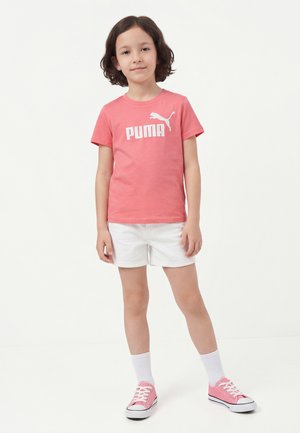 Copil în picioare, îmbrăcat într-un tricou Puma coral, pantaloni scurți albi, șosete albe și adidași roz pe un fundal alb.