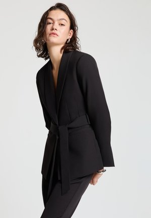 Junge Frau mit kurzen dunklen Haaren, die einen schwarzen Blazer mit Gürtel und schwarze Hosen trägt, posiert vor einem schlichten hellen Hintergrund.