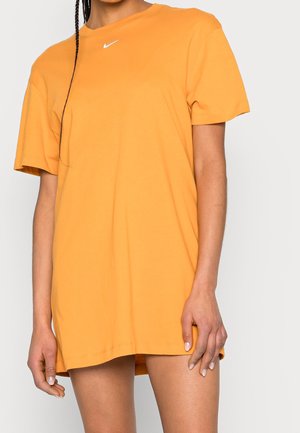 Person iført løs, kortærmet orange t-shirt kjole med lille hvid Nike-logo nær halsudskæringen, stående mod ensfarvet baggrund.