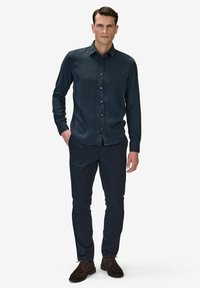 Mörkblå skjorta med knappar i ett smooth tyg med krage, kombinerad med mörka byxor och bruna mockaskor. Stående hållning, neutral bakgrund.