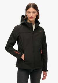 Zwarte softshell jas met een capuchon, ritssluiting aan de voorkant, contrasterende oranje accenten en een aansluitende pasvorm. Voorzien van zijzakken en elastische manchetten.