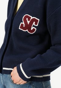 Ναυτικό κάρντιγκαν από πλεκτό ύφασμα με υφή, διαθέτοντας μάρκας "SC" σε σκούρο κόκκινο και λευκές ρίγες στο μανίκι και το στρίφωμα.