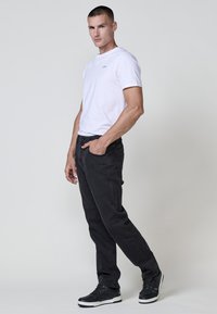 Camiseta blanca de manga corta, jeans de mezclilla negros, tela texturizada y zapatillas deportivas negras con acentos blancos. El modelo está en una pose relajada.