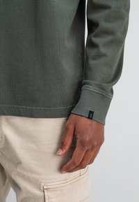 Close-up van de rechterhand van een persoon met een zichtbare litteken, gekleed in een olijfgroen lange mouw shirt en beige cargopantalon.