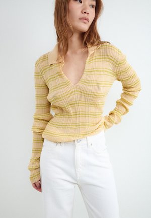 Junge Frau trägt einen gelb-grün gestreiften, gerippten Pullover mit Kragen und weiße, hoch taillierte Hosen, schaut zur Seite.