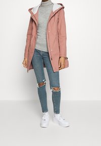 Różowa kurtka z kapturem w szarym wykończeniu, zestawiona z szarym golfem, zniszczonymi niebieskimi dżinsami i białymi sneakersami.