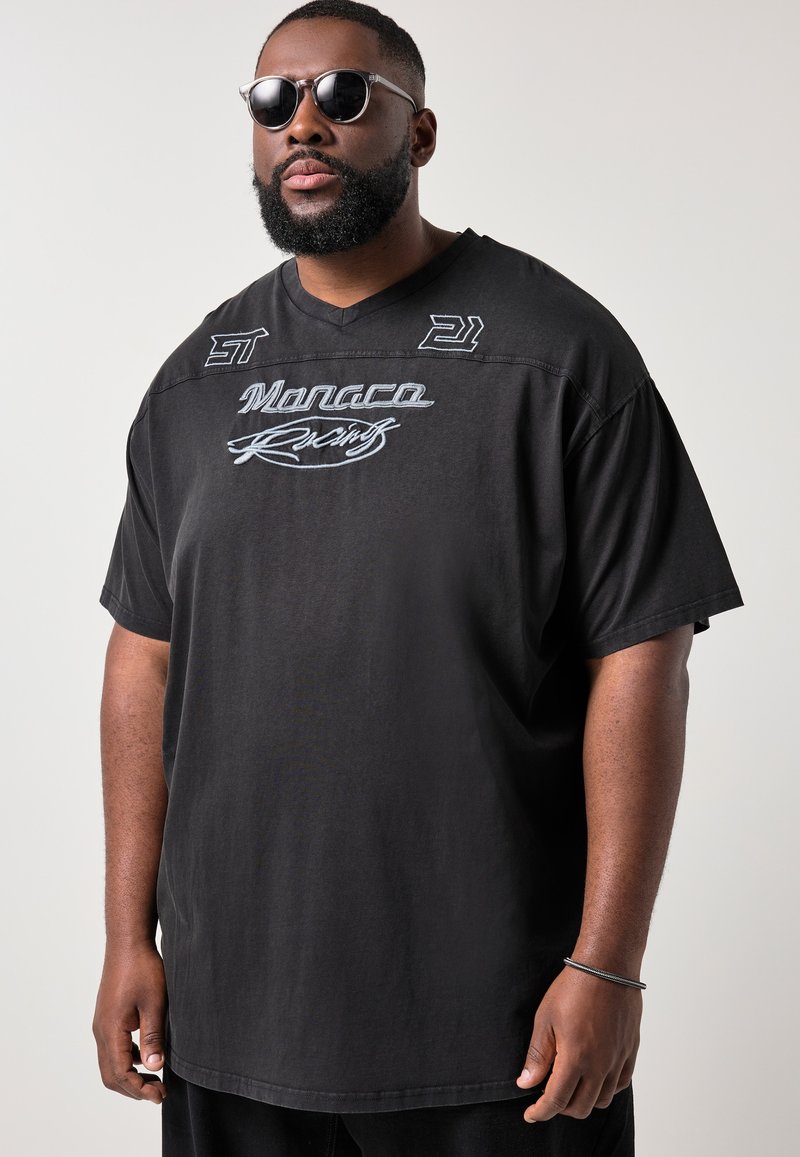 Hombre con barba, lleva una camiseta negra con la inscripción "Monaco Racing" y gafas de sol, posando frente a un fondo claro y liso.