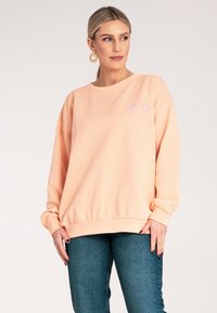 Brzoskwiniowy sweatshirt z okrągłym dekoltem, długimi rękawami i prążkowanym wykończeniem. Na piersi haftowany napis w jasnofioletowym kolorze.