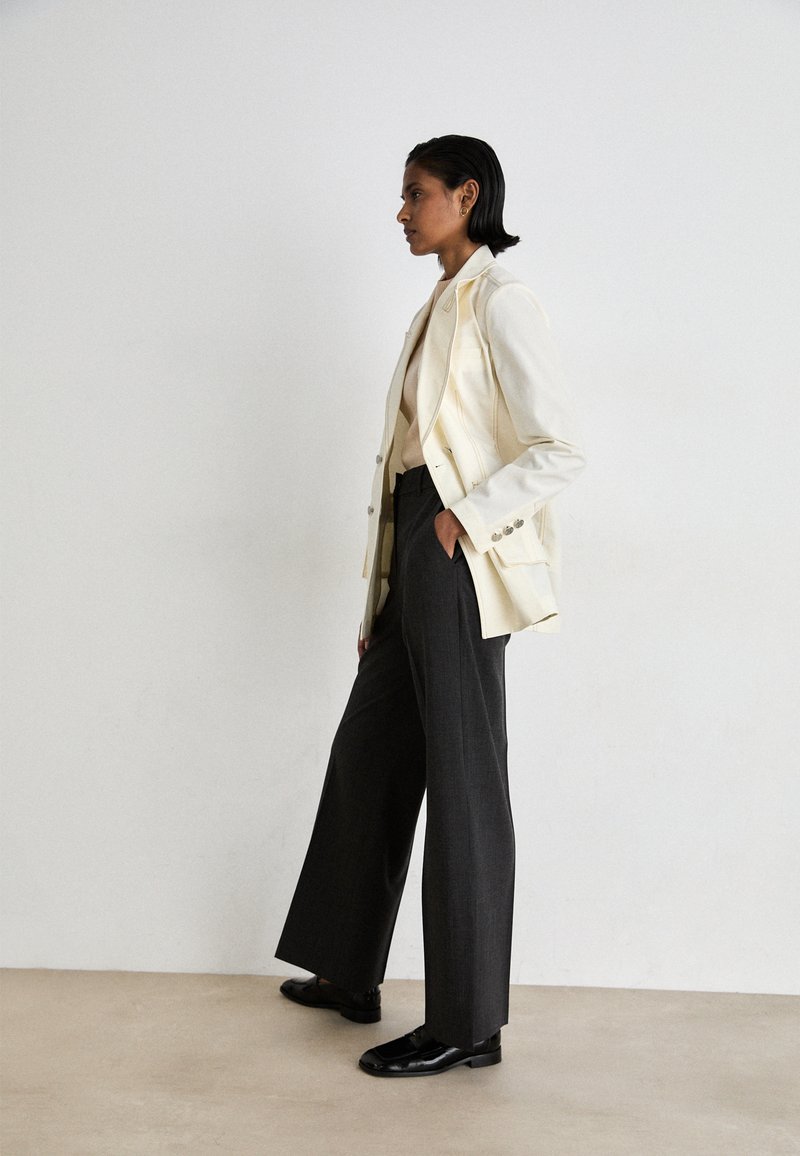 Cremefarbener, doppelt geknöpfter Weste mit Reverskragen, kombiniert mit hoch taillierten, weit geschnittenen grauen Hosen und schwarzen, polierten Schuhen.