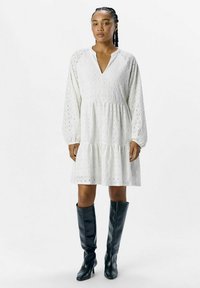 Witte, knielange jurk met een gelaagde rok, lange mouwen, een V-hals en een oogjespatroon. Gepaard met zwarte leren laarzen tot de knie.