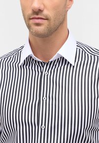 Camicia a righe bianche e nere con colletto classico, linee verticali e bottoni. Il tessuto ha una texture liscia e una vestibilità tailored.