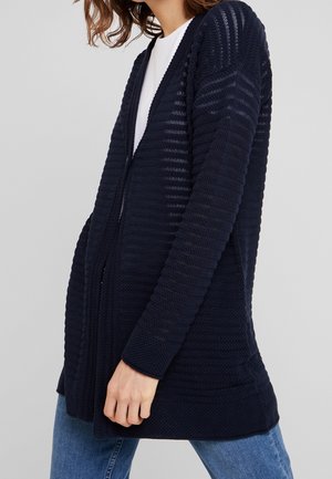 Frau trägt eine marineblaue, offene Strickjacke über einem weißen Top und blauen Jeans vor einem schlichten hellgrauen Hintergrund.