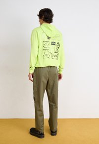 Νεον πράσινη κουκούλα με γραφικά σχέδια, συνδυασμένη με ελαιόχρωμα cargo παντελόνια και μαύρη υπόδηση. Καθημερινός σχεδιασμός, άνετη εφαρμογή και ρυθμιζόμενη κουκούλα.