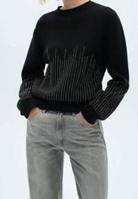 Schwarzer Strickpullover mit Rippentextur, verziert mit silberfarbenen metallischen Details in vertikalen Linien, kombiniert mit hellgrauen Jeans.