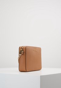 Tan lædertaske med firkantet form, guld kædetræk og lynlåslukning. Har en logotavle på forsiden og struktureret overflade.