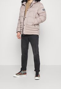 Kurtka puchowa w jasnym beżu z odpinanym kapturem wykończonym czarnym futrem, suwak z przodu i elastyczne mankiety. Nosi ją w połączeniu z czarnymi dżinsami i wzorzystymi sneakersami.